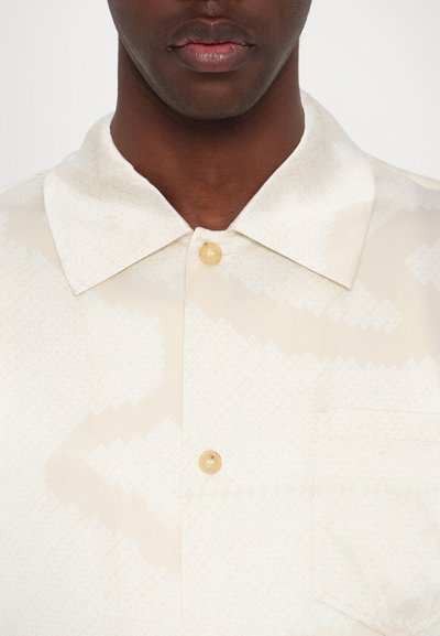 Camisa de manga corta con cuello, hecha de un tejido suave, de color crema con un sutil patrón geométrico, que presenta un bolsillo en el pecho y un botón claro.