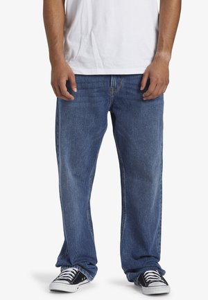 Homme portant un jean bleu ample, des baskets noires et blanches et un t-shirt blanc, debout sur un fond blanc uni.