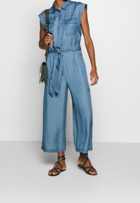 Salopette in denim con design senza maniche, colletto, tasche laterali e vita con cintura. Taglio a gamba larga in azzurro chiaro, abbinata a sandali neri.