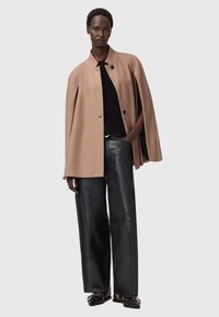 Cappotto in misto lana color cammello con colletto alto e chiusura con bottoni frontali, abbinato a pantaloni in pelle neri a vita alta e scarpe basse.