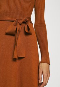 Rostfärgad stickad klänning med ribbat mönster, med långa ärmar och en knytning i midjan som skapar en smidig silhuett.