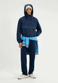 Marineblauer Sportanzug mit einem Kapuzenoberteil mit Viertelreißverschluss und Logo sowie passenden Hosen. Hellblauer Pullover lässig um die Taille gebunden.