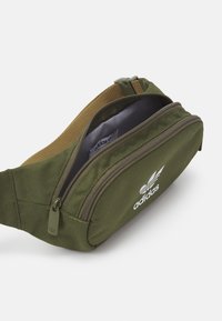 Grön tygmidjeväska med en ficka med dragkedja, med en slät yta, tan detaljer och vit Adidas-logotyp.