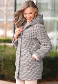 Grau gemusterter Mantel mit Kapuze, aus strukturiertem Stoff, mit vorderen Taschen und Knopfverschluss. Getragen über hellfarbener Bekleidung.