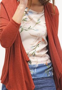 Kardigan w kolorze rdzy, lekki, nałożony na top w kwiatowy wzór w delikatnych pastelowych odcieniach, w zestawieniu z dżinsami. Brak widocznych dodatków lub akcentów.