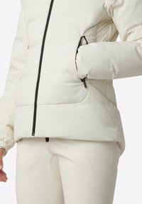 Biała puchowa kurtka z zapinaniem na zamek z przodu, bocznymi kieszeniami i elastycznymi mankietami. Miękka, gładka tkanina o matowym wykończeniu, zestawiona z jasnymi spodniami.