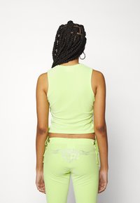 Femme aux longs cheveux tressés portant un débardeur vert citron vert et un pantalon assorti avec un motif cœur en strass sur les poches arrière.