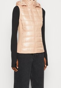 Padded vest w kolorze jasnoróżowym, z błyszczącym wykończeniem, na zamek błyskawiczny i z kapturem. Czarna koszula z długim rękawem pod ciemnymi dżinsami.