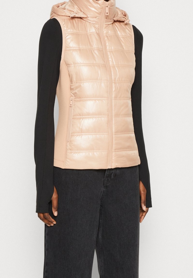 Padded vest w kolorze jasnoróżowym, z błyszczącym wykończeniem, na zamek błyskawiczny i z kapturem. Czarna koszula z długim rękawem pod ciemnymi dżinsami.