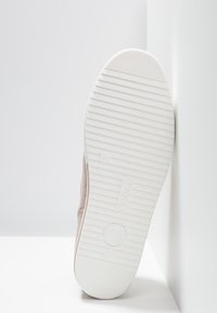 Biała gumowa podeszwa z teksturowanymi rowkami; jasnobeżowy materiał górny; mały detal marki z boku i gładkie wykończenie.