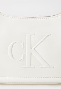 Valge nahast käekott, millel on reljeefne 'CK' logo. Kotil on sujuv tekstuur, ümarad servad ja peen tõmblukk detail ülaosas.