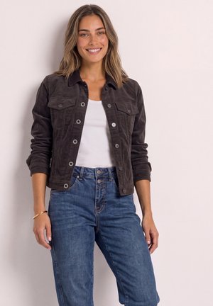 Junge Frau lächelt, trägt eine dunkle, geknöpfte Jacke, weißes Oberteil, blaue hochgeschnittene Jeans, mit offenem Haar und einem goldenen Armband am linken Handgelenk.