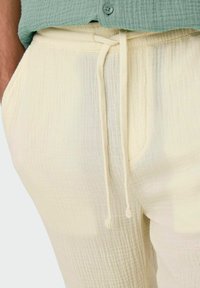 Pantalons texturés de couleur crème avec une taille élastique et un cordon de serrage. Dotés de poches latérales et d'une coupe décontractée, portés avec un haut vert.