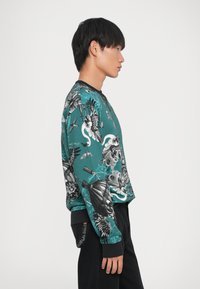 Felpa verde a maniche lunghe con intricate grafiche in bianco e nero, che presenta motivi di uccelli, elementi floreali e fantasie fantasiose.