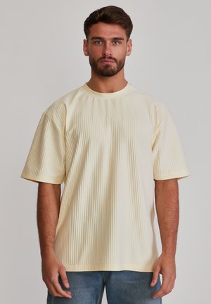 Hombre con cabello corto y oscuro y barba, vestido con una camiseta crema de manga corta acanalada y vaqueros azules, de pie frente a un fondo liso.