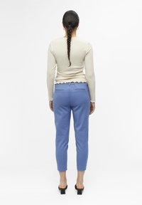 Jasno beżowa bluzka z długim rękawem o prążkowanej fakturze; niebieskie zwężane spodnie z elastycznym paskiem w talii i falbanką na końcach; przedstawiony widok z tyłu.