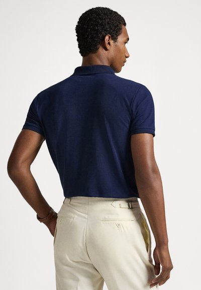Μπλε ναυτικό πόλο πουκάμισο από απαλό ύφασμα, με κοντά μανίκια και κλασικό γιακά. Συνδυάζεται με ανοιχτό μπεζ παντελόνια με θηλιές για ζώνη και τσέπες.