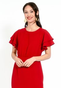 Rochie roșie cu mâneci scurte și volănașe, talie ajustată, material neted și decolteu rotund. Model purtând cercei mari din aur, zâmbind.