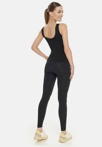 Črn top brez rokavov z širokimi naramnicami, prilegajočega kroja in zaobljenim hrbtom, v kombinaciji s črnimi leggingsi. Model nosi svetle športne copate.