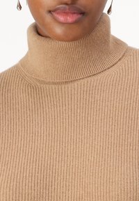 Pull à col roulé beige côtelé en tissu tricoté. Comprend un col haut et un motif texturé, mettant en avant la chaleur et le confort.