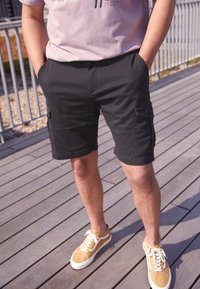 Pantalones cortos cargo negros con bolsillos laterales, combinados con zapatillas beige con detalles blancos, sobre una superficie de madera.