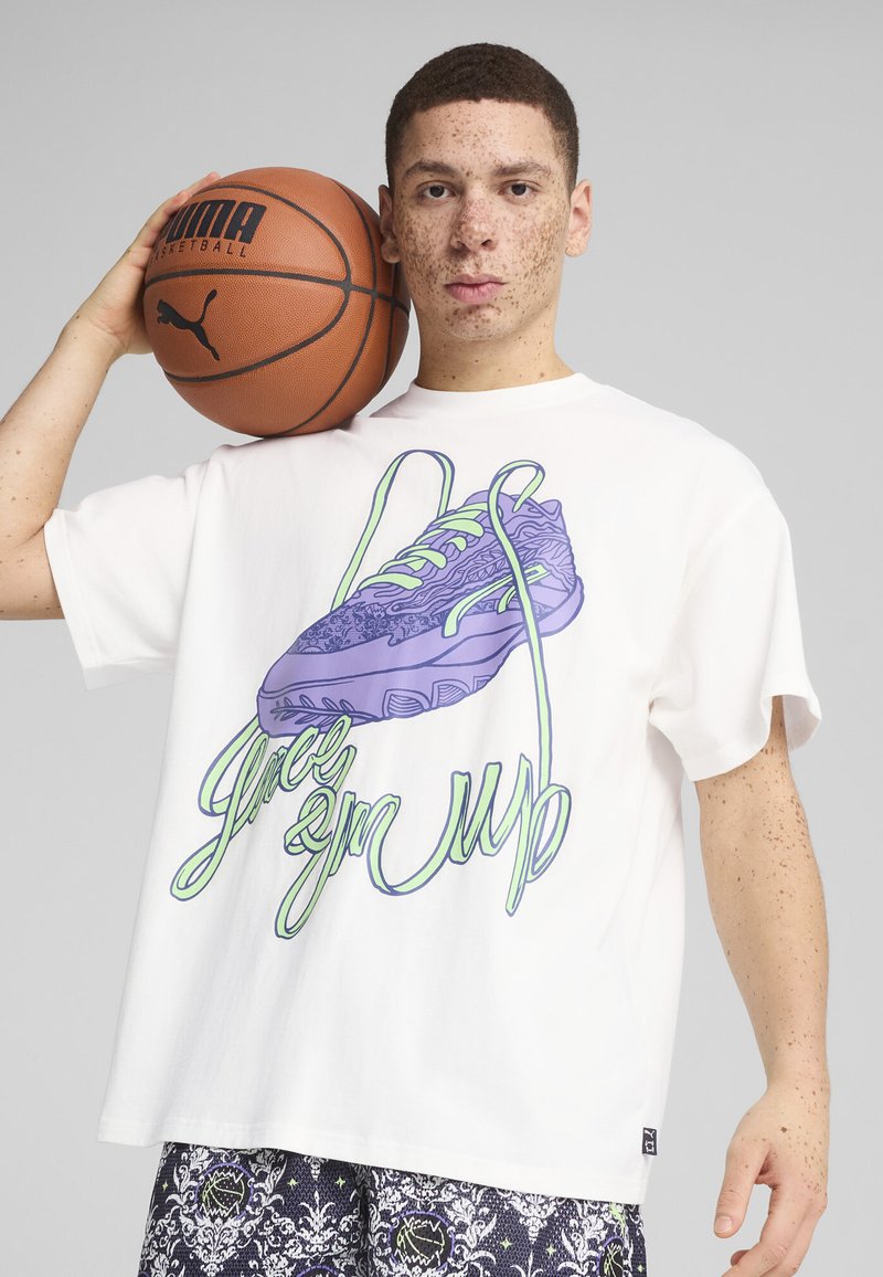 Bílý bavlněný tričko s grafickým vyobrazením purpurového tenisky se zelenými tkaničkami a textem "zavázat je". Model drží basketbal a má na sobě vzorované šortky.