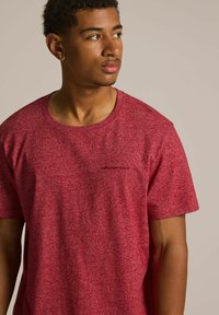 Czerwona koszulka z krótkim rękawem w kropki, z okrągłym dekoltem i małym czarnym logo na klatce piersiowej. Materiał bawełniany lub mieszanka bawełny.