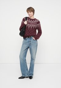Bordeaux gebreide trui met "Vrolijk Kerstmis" print, blauwe uitlopende jeans, zwarte tas en zwarte puntige schoenen. Gladde textuur, ontspannen pasvorm.