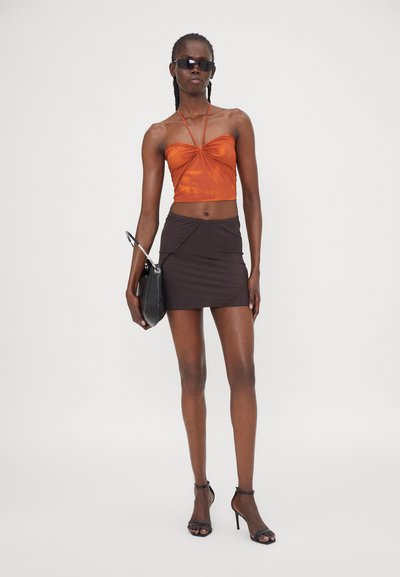 Top court en satin orange avec un devant froncé, associé à une mini-jupe marron foncé à design asymétrique. Les accessoires comprennent des lunettes de soleil noires et un petit sac à main.