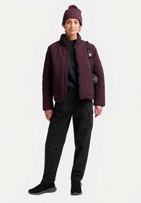 Giacca imbottita bordeaux con motivo trapuntato, abbinata a pantaloni cargo neri e un berretto lavorato a maglia. Scarpe nere completano il look.