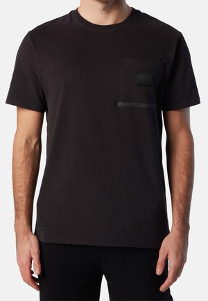 Uomo che indossa una t-shirt nera a maniche corte semplice con una piccola tasca a toppa nera sul lato sinistro del petto e sottili righe orizzontali appena visibili.