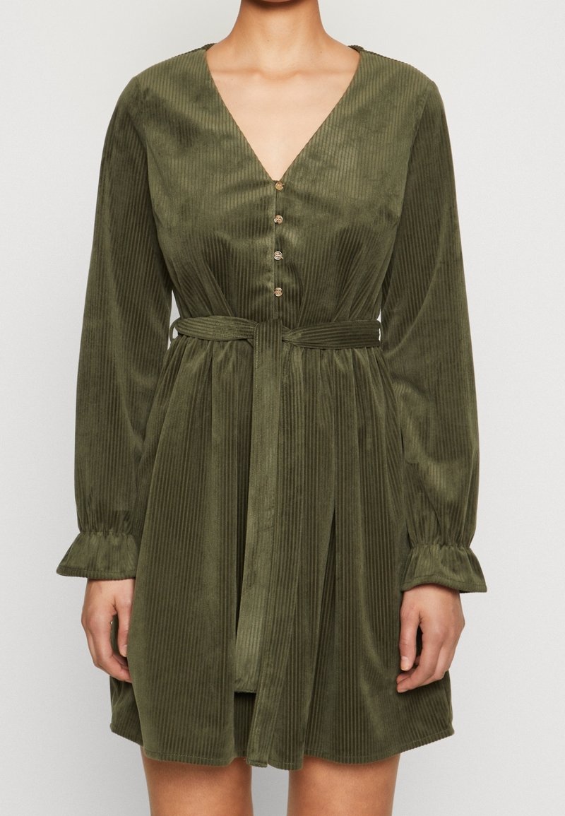 Femme portant une robe ceinturée verte olive à manches longues avec une texture côtelée verticale et un col en V boutonné sur le devant.