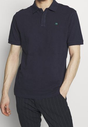 Mand iført en ensfarvet mørk navy poloshirt med en lille grøn symaskinelogo på brystet, kombineret med mørke bukser med knapper.