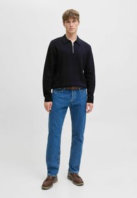 Jauns vīrietis, tērpts melnā garajā piedurknē ar rāvējslēdzēju, zili džinsi, brūns jostas un brūnas kurpes, stāv uz vienkārša balta fona.