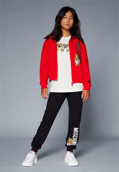 Rød zip-up hettegenser, hvit t-skjorte med tegneserie-graphic, svarte joggebukser med logo, og hvite sneakers. Casual, komfortabel antrekk.