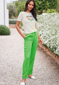 Light gray t-shirt with colorful "BEACH" graphic, paired with vibrant green linen pants and white slide sandals, set in a garden backdrop.