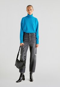 Helle blaue Rollkragenpullover, hochtaillierte graue Jeans mit Knöpfen, schwarze Schultertasche mit Nieten und glänzende schwarze Stiefeletten.