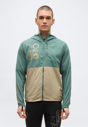 Vyras dėvi žalią ir smėlinę striukę su užtrauktuku, su apskritais grafikais ant krūtinės ir juodus kelnes su baltais linijinių piešinių ir teksto elementais.