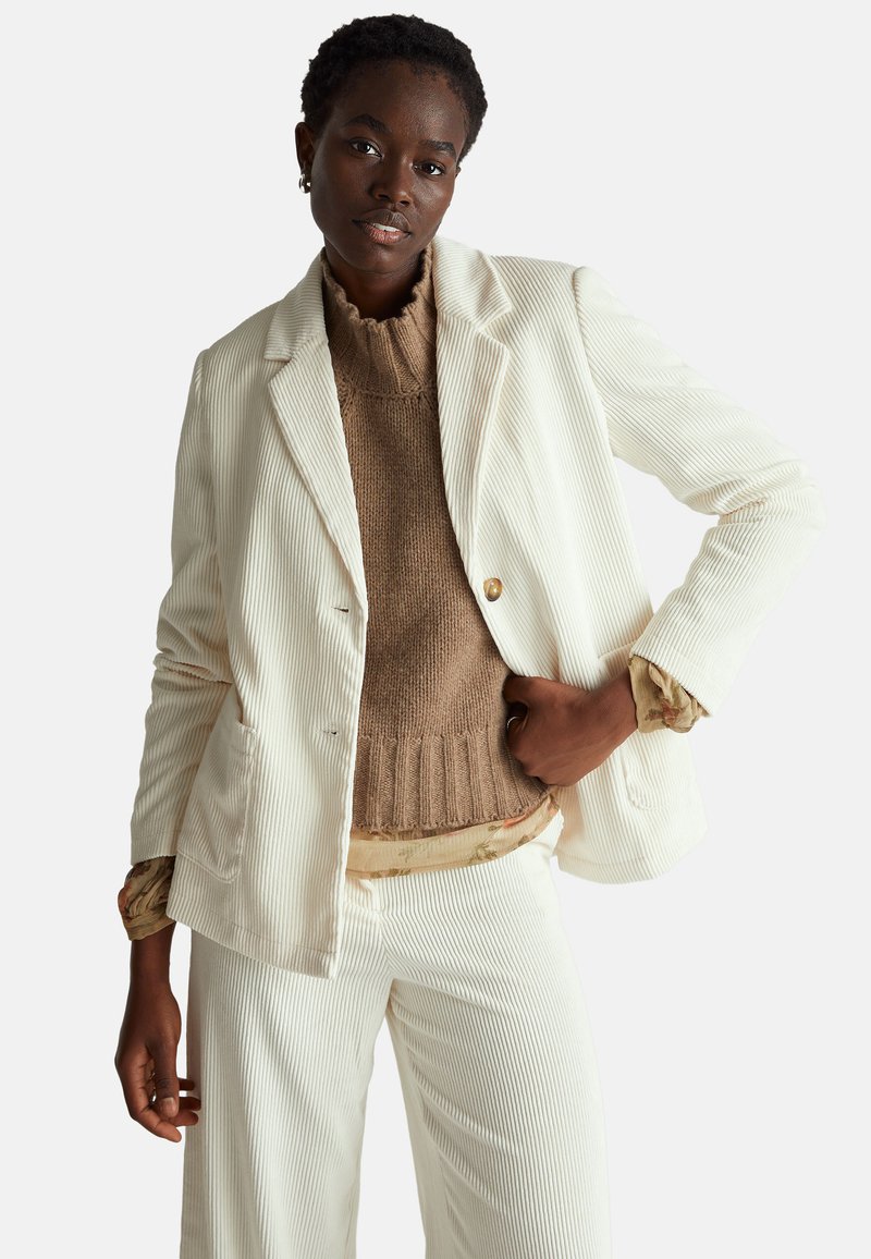 Dvoudílný outfit obsahující bílý kordový blazer s vertikálními pruhy, hnědý pletený svetr a odpovídající široké kordové kalhoty.