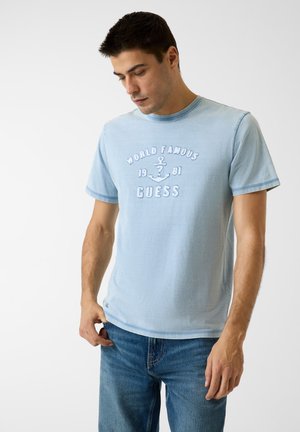 Hellblaues Baumwoll-T-Shirt mit Rundhalsausschnitt, kurzen Ärmeln und besticktem Schriftzug "WORLD FAMOUS 1981 GUESS" sowie Anker-Design auf der Vorderseite.