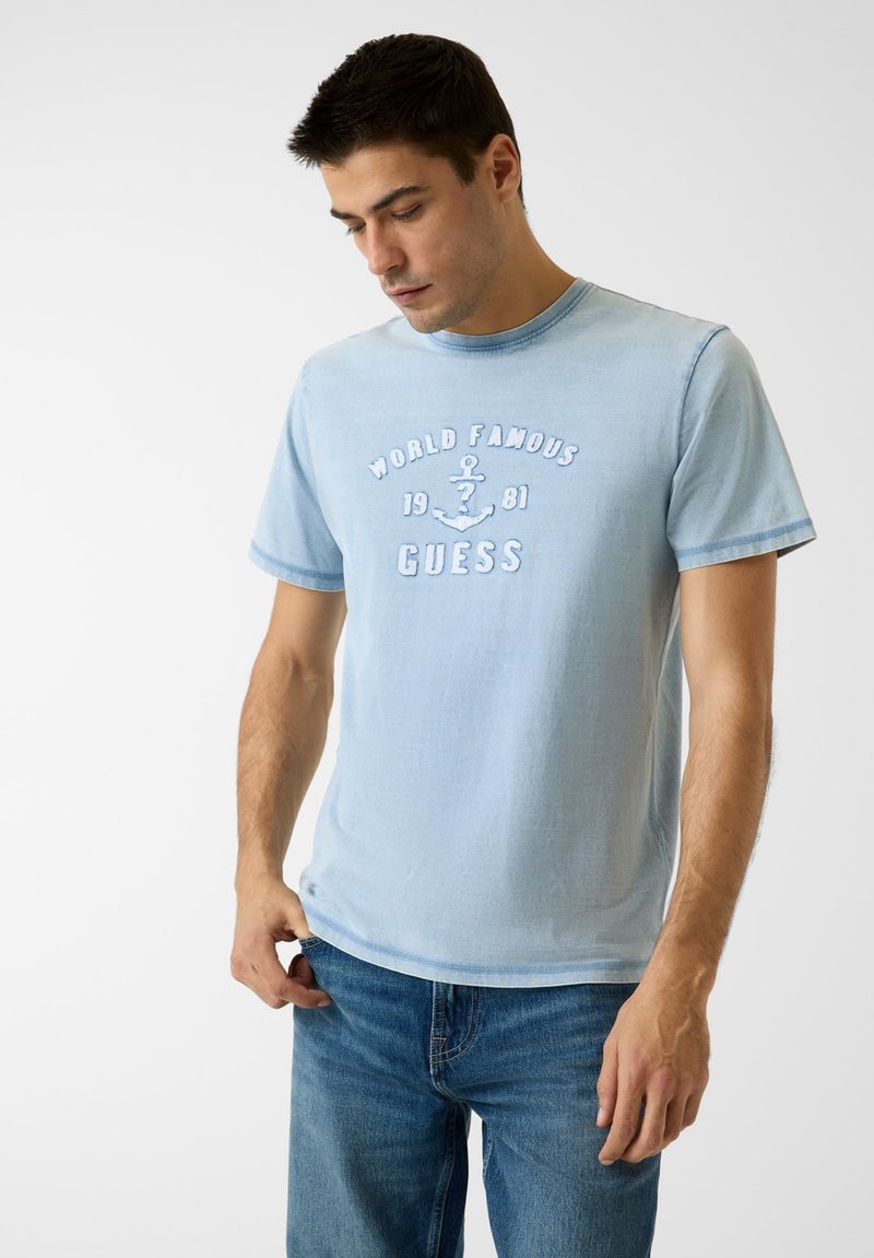 T-shirt en coton bleu clair avec un col rond, des manches courtes, et une broderie "WORLD FAMOUS 1981 GUESS" accompagnée d'un motif d'ancre sur le devant.