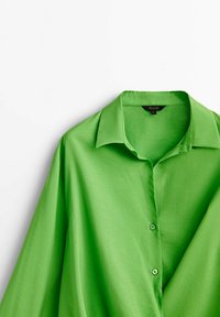 Blouse verte à manches longues avec un col, des boutons à l'avant et une texture de tissu lisse. Légère, avec une coupe ample.