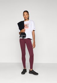 White graphic t-shirt with orange print, paired with maroon leggings and black athletic shoes. Model holds a black towel over her shoulder.