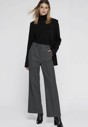 Donna con dolcevita nero, blazer nero, pantaloni grigi a gamba larga e stivaletti neri con tacco, in piedi con una mano in tasca.