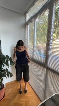 Femme ajustant un short noir ample debout près d'une grande fenêtre avec des stores, à côté d'une plante verte en pot sur un sol en bois.