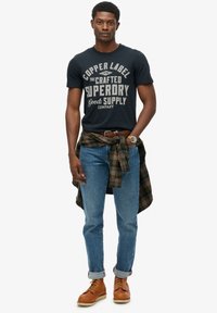 Marineblauw grafisch T-shirt met 'Superdry' opdruk, blauwe spijkerbroek en bruine laarzen. Groene geruite blouse in de taille geknoopt. Horloge om de pols.