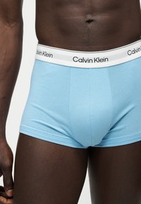 Άνδρας που φορά ανοιχτό μπλε Calvin Klein μποξεράκια με λευκή ζώνη μέσης και μαύρο λογότυπο, εμφανιζόμενος από τη μέση έως το μέσο του μηρού.