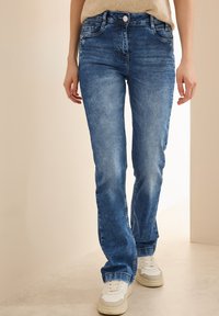 Blå denimjeans med rak skärning, ljus blekning, framfickor och kn�äppning, kombinerade med vita sneakers mot en neutral bakgrund.