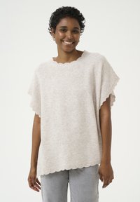 Licht beige gebreide trui met een golvende halslijn en korte mouwen, met een relaxte pasvorm en een zachte textuur. Gecombineerd met grijze jeans.