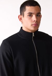 Čierna zateplená bunda na zips s vysokým golierom, vyrobená z hladkej látky. Obsahuje kovový zips a rebrovanú textúru na golieri.